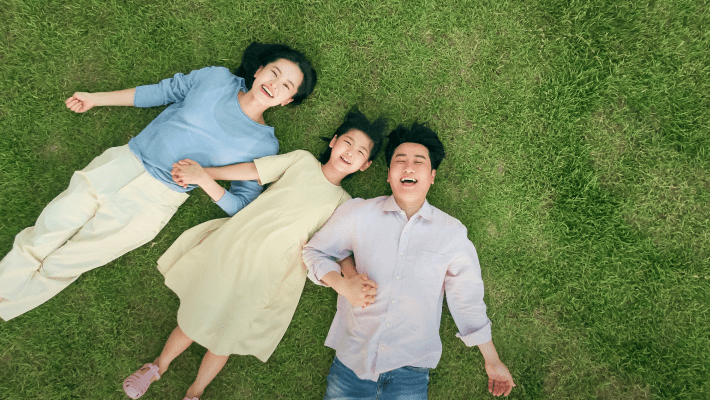 초록의 잔디밭에 어린 아이를 포함한 세가족이 나란히 손을 잡고 누워서 밝게 웃고 있는 이미지 입니다.